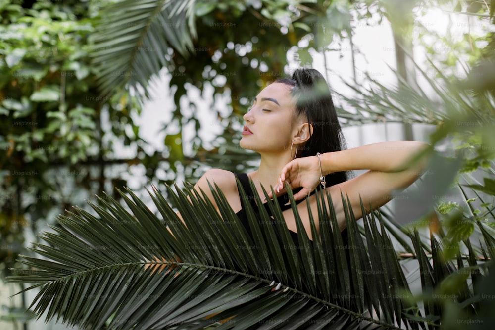 Beautiful mixed raced Asian woman model with dark long ponytail hair style, wearing black dress, posing in greenhouse with tropical plants, standing behind palm tree leaf. Tropical, summer concept