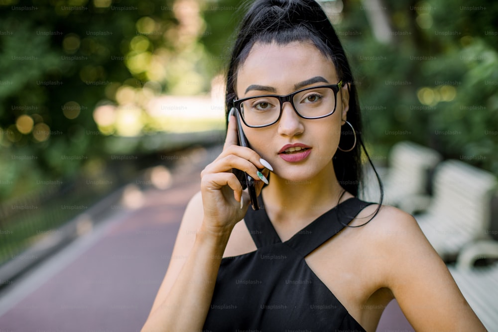 Lifestyle close up portrait of beautiful confident Asian business woman in park outdoors, wearing black dress and eyeglasses, talking by smart phone, standing over city park background.