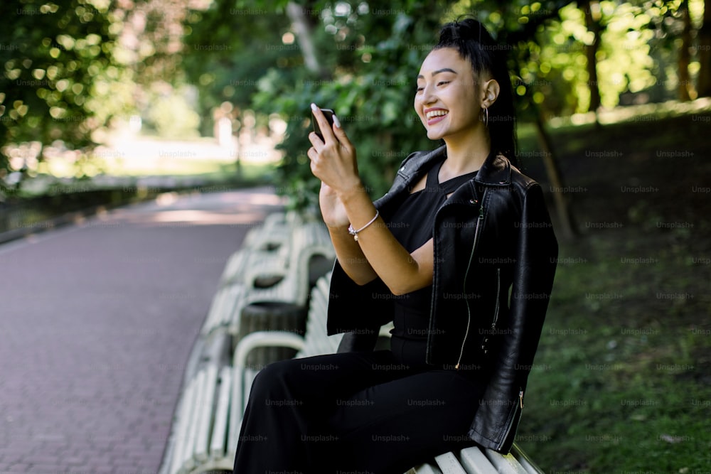 Schöne fröhliche junge asiatische brünette Frau, die trendiges schwarzes Outfit trägt, auf der Bank im Stadtpark sitzt, ihr Handy hält, während sie lustige Memes oder Nachrichten liest.