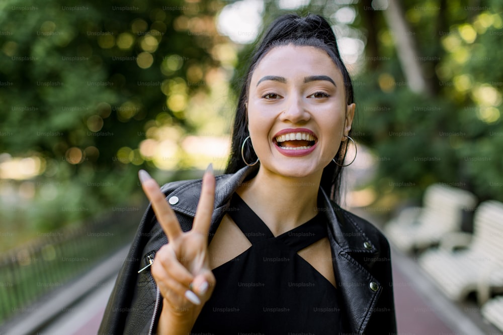 Horizontal outdoor portrait of beautiful laughing Asian young woman, wearing black leather jacket, smiling to camera and showing fingers doing victory peace sign. Asian lady in city park.