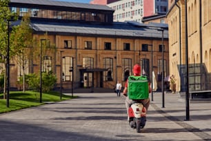 Vista trasera del mensajero con bolsa de refrigerador montando scooter a lo largo de una hermosa calle soleada con pequeñas casas. Concepto de entrega de alimentos