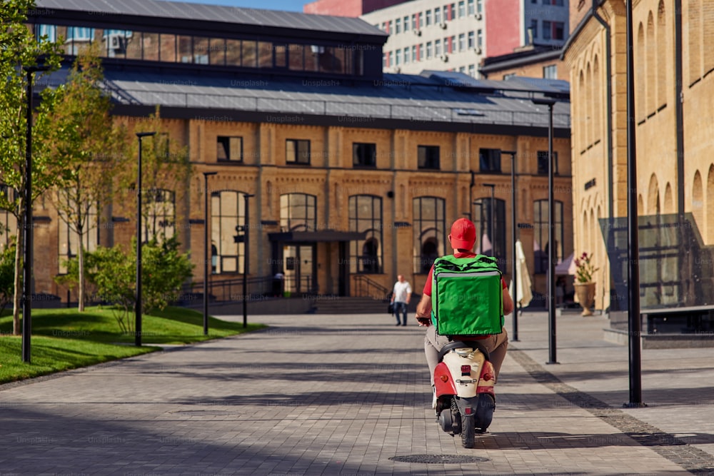 Rear view of courier with refrigerator bag riding scooter along beautiful sunny street with small houses. Food delivery concept