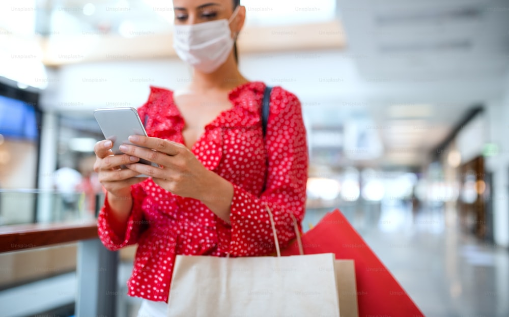 Woman with face mask standing with smartphone indoors in shopping center, coronavirus concept. Copy space.