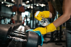 Young female worker disinfecting cleaning and weeping expensive fitness gym equipment with alcohol sprayer and cloth. Coronavirus global world pandemic and health protection safety measures.