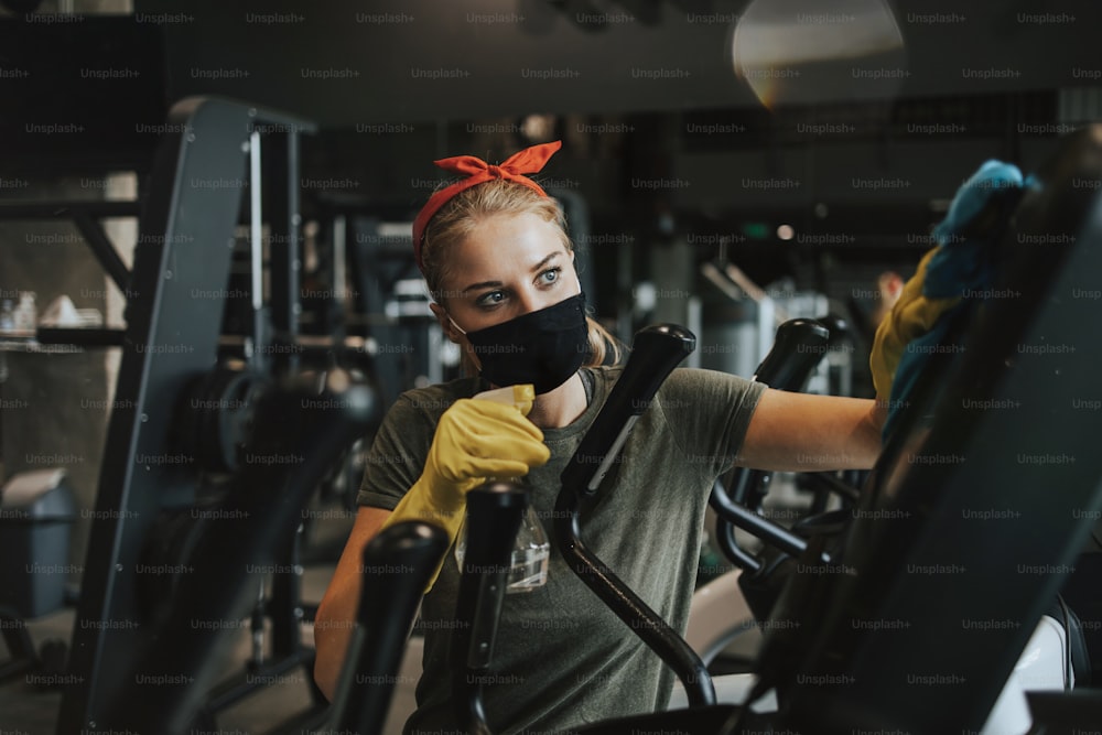 Young female worker disinfecting cleaning and weeping expensive fitness gym equipment with alcohol sprayer and cloth. Coronavirus global world pandemic and health protection safety measures.