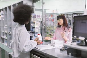 Back angle view of female Afrcian pharmacist giving medicine to young smiling Caucasian woman in drugstore. Pharmacist dispensing drug to patient in pharmacy.