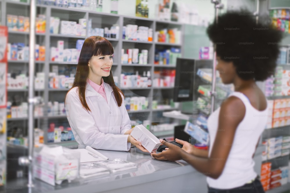Young pretty Caucasian woman pharmacist helping a female African customer, dispensing her prescription medicine with a friendly smile. Pharmacist and client at modern drugstore.