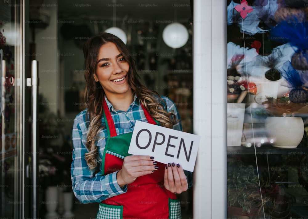 Young happy woman working in city street flower shop or florist. She is holding open sign while standing on shop doors and smiling. Open for business concept.