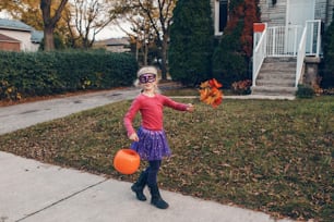 Des bonbons ou des bâtons. Fille enfant heureuse avec un panier de citrouille rouge allant à la chasse aux bonbons pendant les vacances d’Halloween. Enfant mignon en costume de sorcière de fête allant chez les voisins pour des bonbons et des friandises.