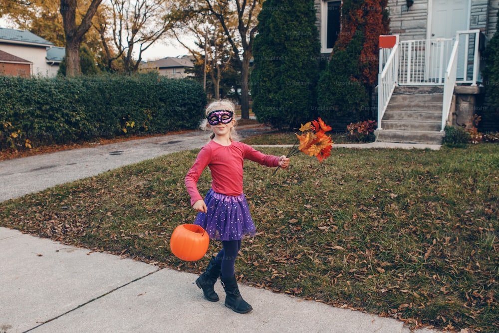 Süßes oder Saures. Glückliches Kindermädchen mit rotem Kürbiskorb, das am Halloween-Feiertag Süßes oder Saures macht. Süßes Kind im Partyhexenkostüm, das zu Nachbarhäusern geht, um Süßigkeiten und Leckereien zu kaufen.