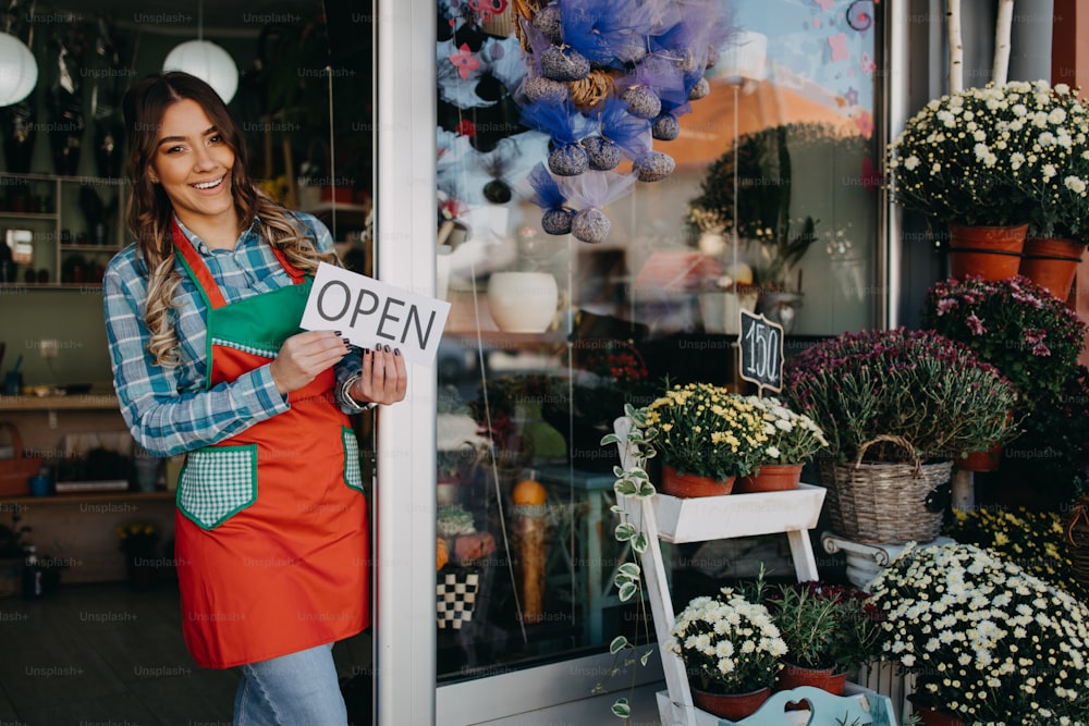 Young happy woman working in city street flower shop or florist. She is holding open sign while standing on shop doors and smiling. Open for business concept.
