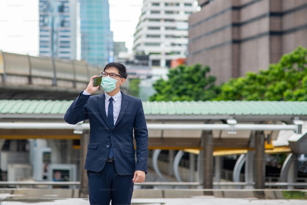 Hombre de negocios asiático con máscara protectora caminando por la ciudad hablando por teléfono