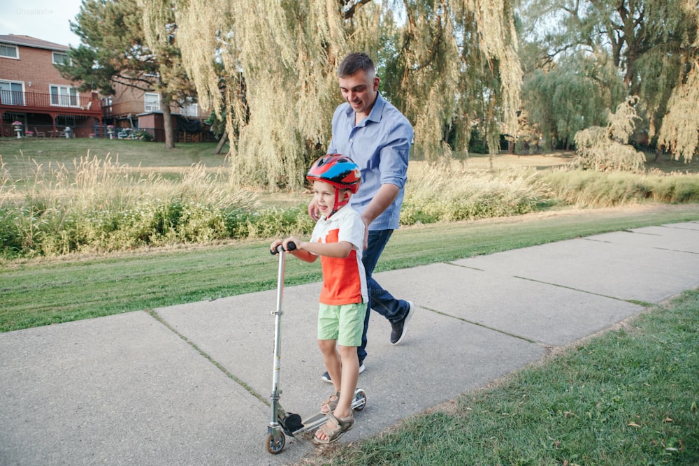 Padre caucásico padre entrenando enseñando a niño hijo a andar en scooter. Niño en edad preescolar con casco con bicicleta en el camino del parque trasero. Actividad familiar infantil de verano de temporada al aire libre.