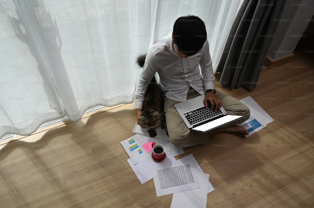 若い男性のフリーランサーは、自宅で猫と一緒に床に座りながらラップトップで作業しています。