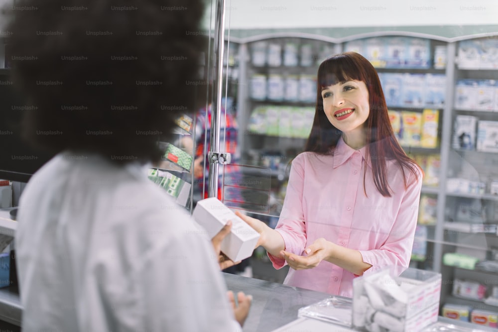 Back view of young african female pharmacist selling medicines to young beautiful Caucasian woman patient. Drug dispensing at modern pharmacy.