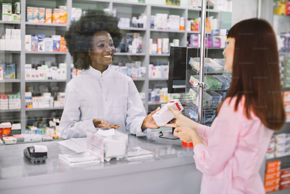 Happy young African female pharmacist standing behind the counter and offering medicine box, talking with young woman customer. Medicine sale, healthcare concept.