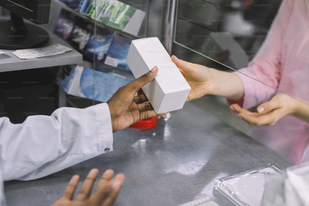 Medicine, pharmaceutics, health care and people concept. Cropped close up image of hands of African female pharmacist giving white box with drug to woman customer at drugstore.