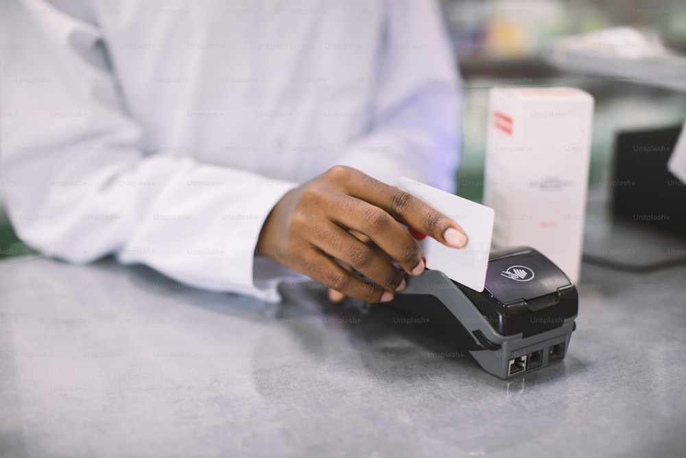 Comprar com cartão de crédito na farmácia. Imagem recortada de mãos de farmacêutica africana segurando cartão de crédito de cliente e um terminal, fazendo pagamento por medicamentos.