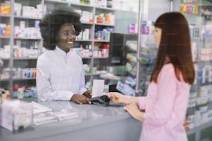 Hübsche junge kaukasische Frau, die für Medikamente mit Kreditkarte in der Apotheke bezahlt. Lächelnde attraktive afrikanische Apothekerin verteilt Medikamente für junge Frau in der Apotheke.
