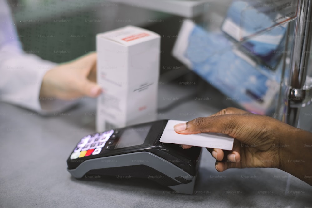Close up cropped image of African woman customer making payment in drugstore with credit card. Pharmacist woman holding white box of drug, woman paying for medicaments.