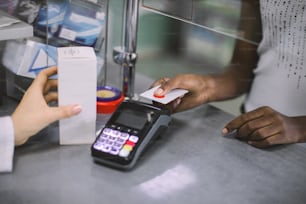Cropped image of hands of female pharmacist holding box of medicine and hand of African woman client patient, paying for medicaments with credit card in pharmacy drugstore