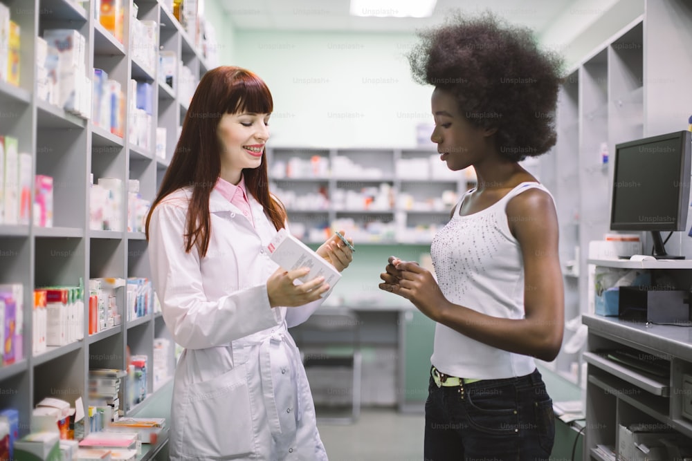 아름다운 전문 여성 약사가 젊은 아프리카 여성 환자와 이야기하면서 두 가지 복용 형태의 약을 보여주고 사용 방법을 설명합니다. 제약 관리.