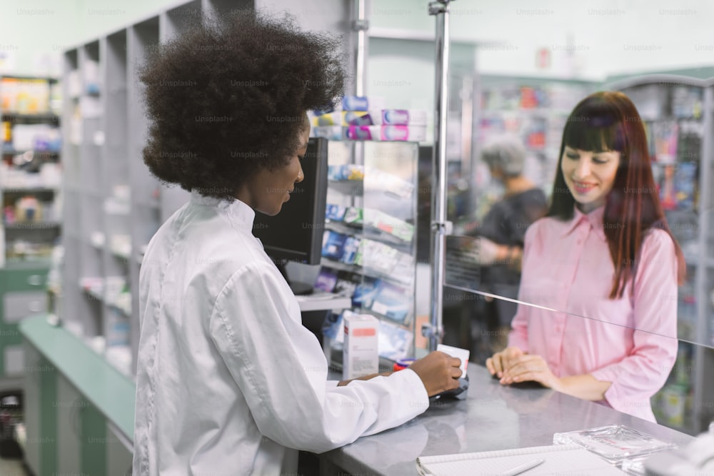 Pretty young Caucasian woman paying for medicines with credit card in modern pharmacy. Back view of African woman pharmacist dispensing drugs for young woman at pharmacy.