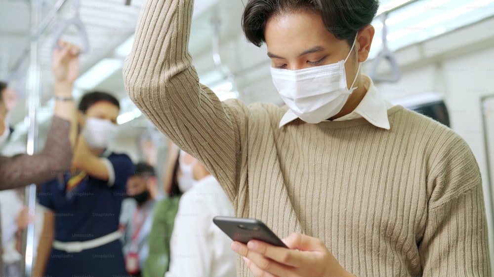 Traveler wearing face mask while using mobile phone on public train . Coronavirus disease or COVID 19 pandemic outbreak and urban city lifestyle problem in rush hour commuting concept .