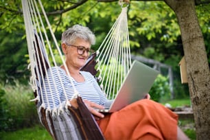Side view portrait of active senior woman with laptop working outdoors in garden, home office concept.