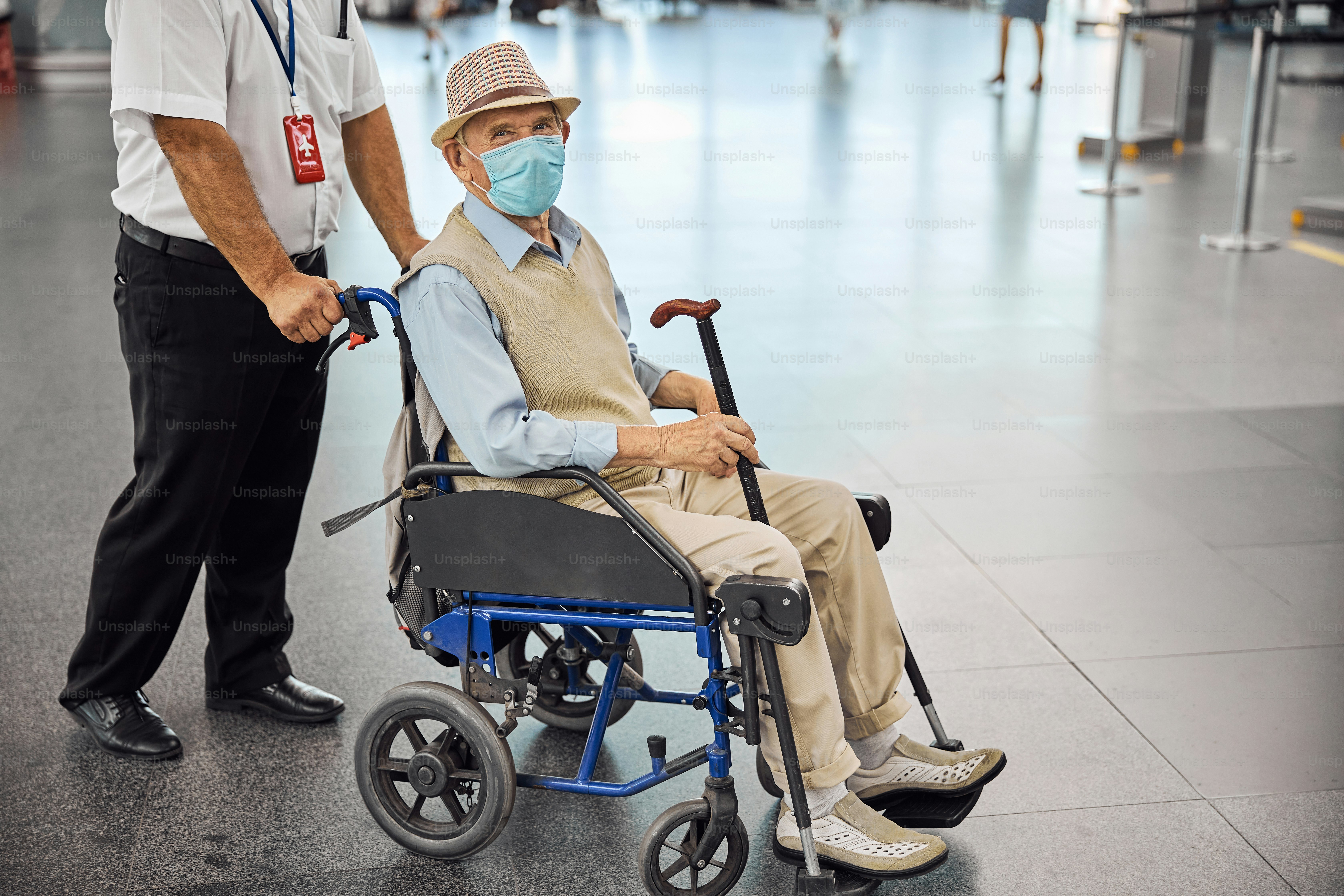 Caucasian airport male employee rolling a transport chair with a senior disabled passenger in a face mask