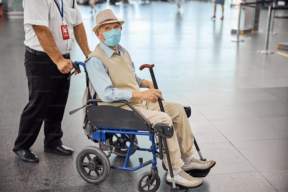Funcionário do aeroporto caucasiano enrolando uma cadeira de transporte com um passageiro com deficiência sênior em uma máscara facial