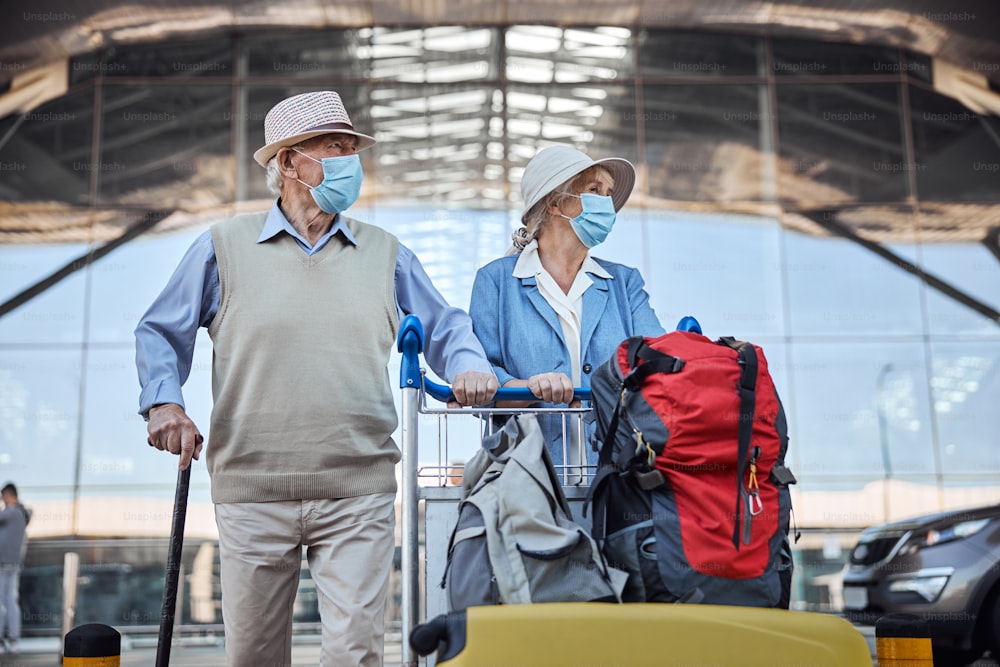 Anciana y su esposo con máscaras faciales y sombreros llevando su carrito de equipaje adelante