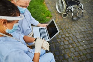 Doctor wearing mask and medical plastic helmet looking at the wristwatch to check the time while sitting near elderly patient