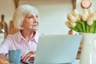 Cintura para arriba de una mujer mayor sonriente sentada en la mesa con una computadora portátil por la mañana mientras mira hacia otro lado. Concepto de estilo de vida doméstico