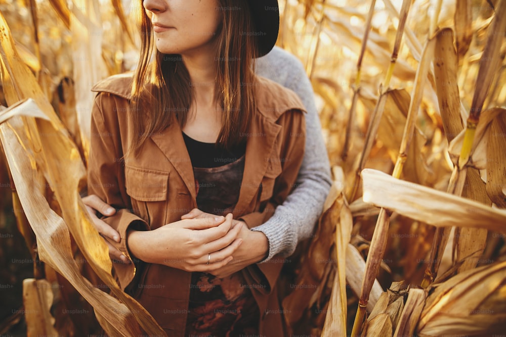 Young man gently hugging his fashionable woman, touch close up among golden corn leaves. Happy stylish couple embracing in autumn corn field in warm sunset light. Romantic sensual moment