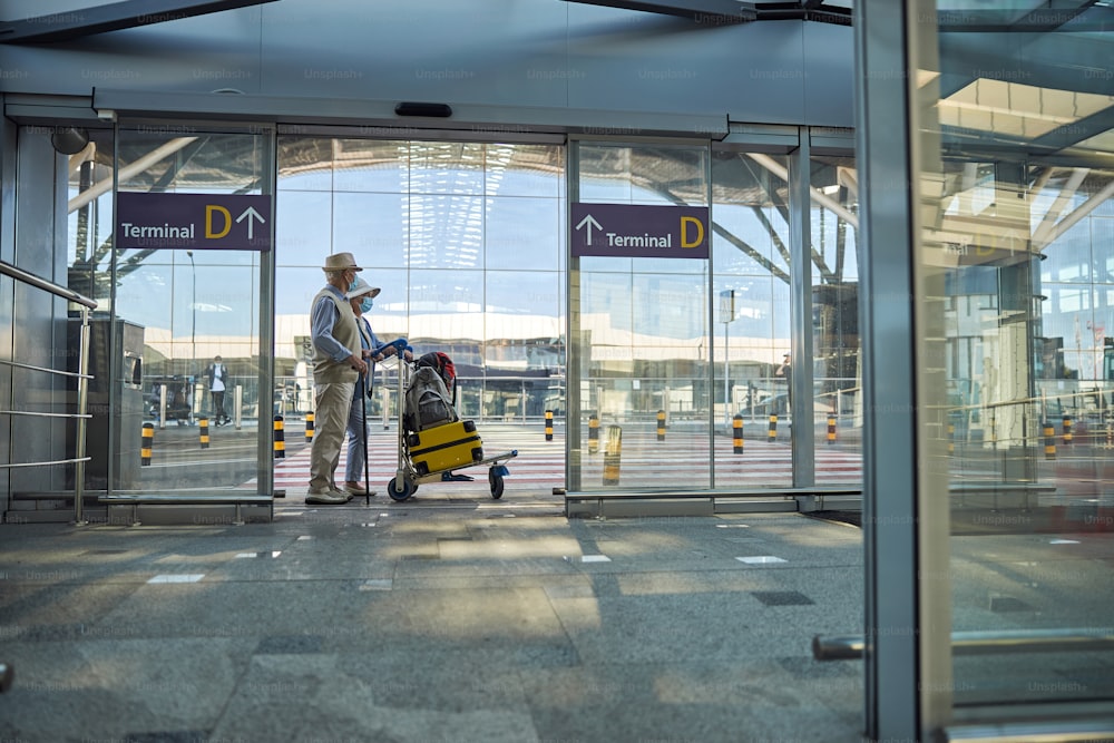 Side view of a senior man in a hat and his wife standing at the terminal entrance