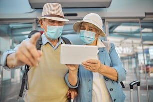 Front view of an elderly tourist couple in face masks staring at the tablet computer