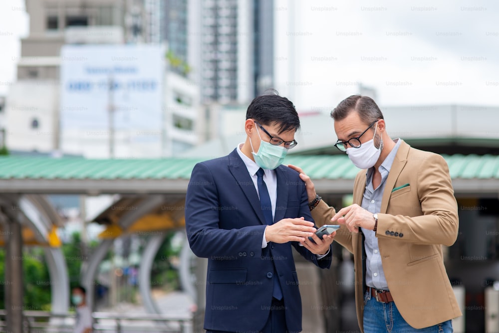 Zwei Geschäftsmann-Kollegen mit Schutzmaske diskutieren Geschäftsprojekt mit Smartphone in der Stadt