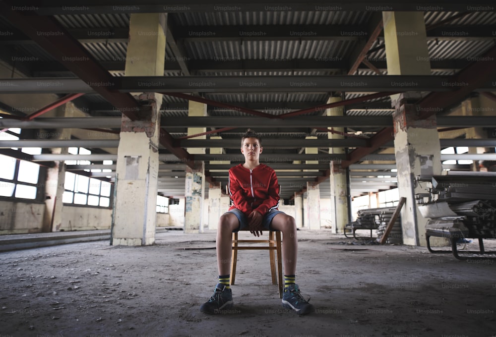 Trauriger und enttäuschter Teenager-Junge, der drinnen in einem verlassenen Gebäude auf einem Stuhl sitzt.