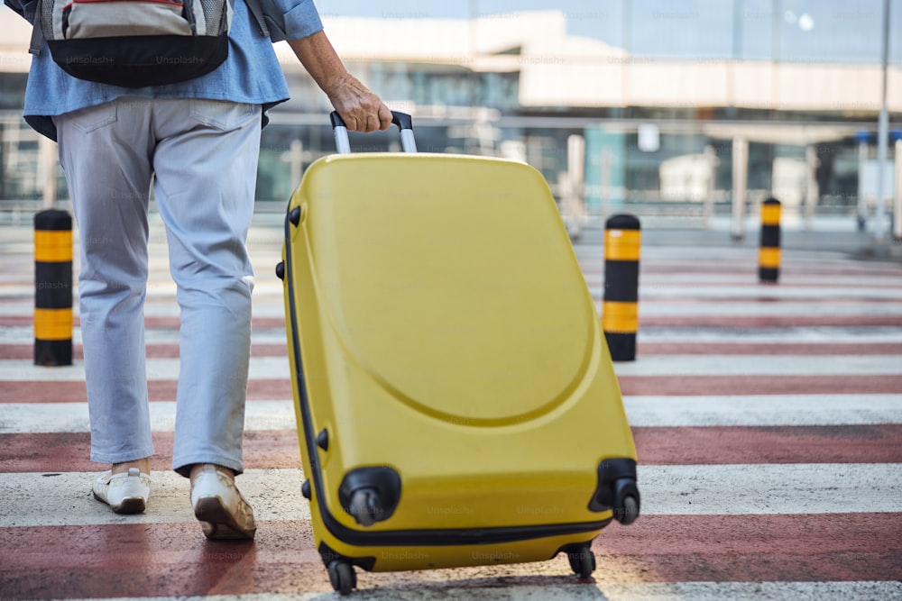 Cropped photo of an elderly Caucasian female tourist in casual clothes wheeling her trolley suitcase