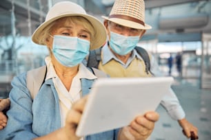 일회용 보호 마스크를 쓴 두 명의 노인 백인 관광객이 태블릿 컴퓨터에서 정보를 검색하고 있다