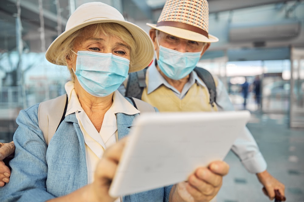 Two aged Caucasian tourists in disposable protective masks searching for information on the tablet computer