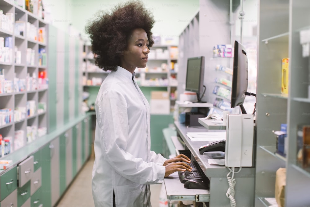 Side view portrait of young confident concentrated African female pharmacist, working with computer behind counter in pharmacy.