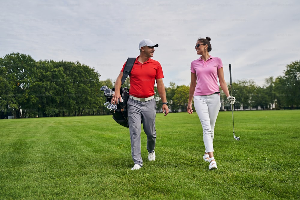 Vista frontal de una dama sonriente con un palo de golf caminando junto a un entrenador profesional