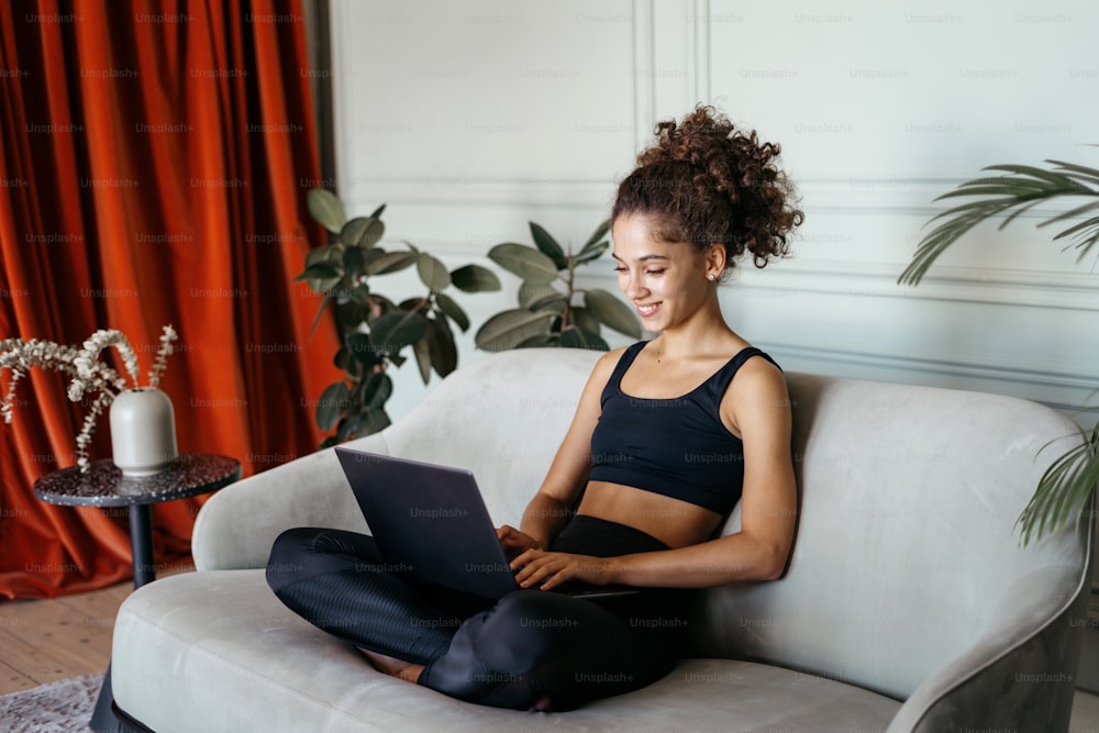 Seitenansicht der lächelnden jungen erwachsenen afro amerikanischen Frau mit modernem Laptop-Computer, sitzt auf Komfortsofa zu Hause im gemütlichen Wohnzimmer und macht glückliches Gesicht. Konzept der Internetkommunikation