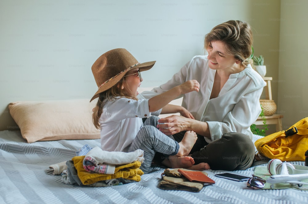 Happy family travel with children concept. Young woman with her daughter having fun packing a backpack for travel sitting on the sofa in the living room.