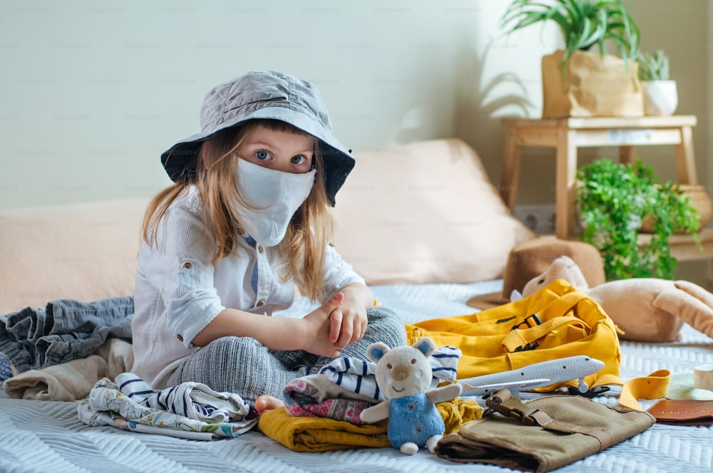 Menina pequena em máscara facial protetora embalando sua mochila para a viagem sentada no sofá dentro de casa. Preparação para uma viagem após o fim da quarentena. Nova vida após a pandemia COVID-19 conceito.