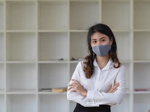 Young confident businesswoman standing with arms crossed in the office room and wearing a face mask