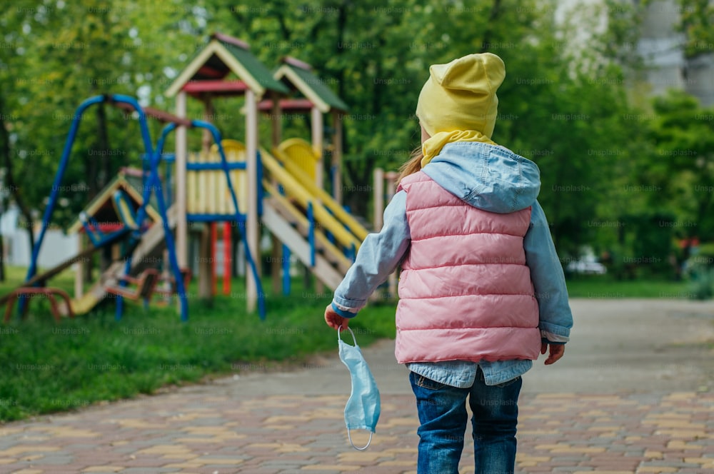 Little girl walking to the kids playground holding protective face mask after the end of quarantine. Pandemic COVID-19 is over concept.