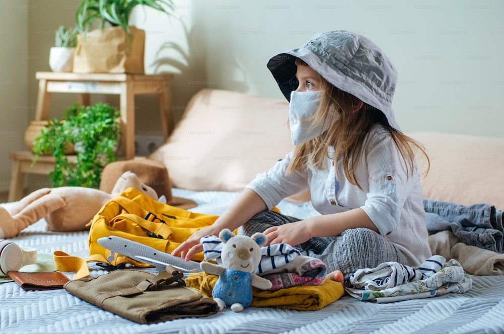 Serious little four years old girl in protective face mask packing her backpack for the travel sitting on the sofa indoor. Prepairing for a travel after the end of quarantine. New life after Pandemic COVID-19 concept.